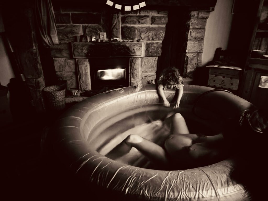 Eine Schwangere liegt im warmen Wasser im Geburtspool, im Hintergrund flackert das Feuer im Kamin und über dem Kaminsims hängen Affirmationskarten zur Motivation: Alleingeburt - ich kann!