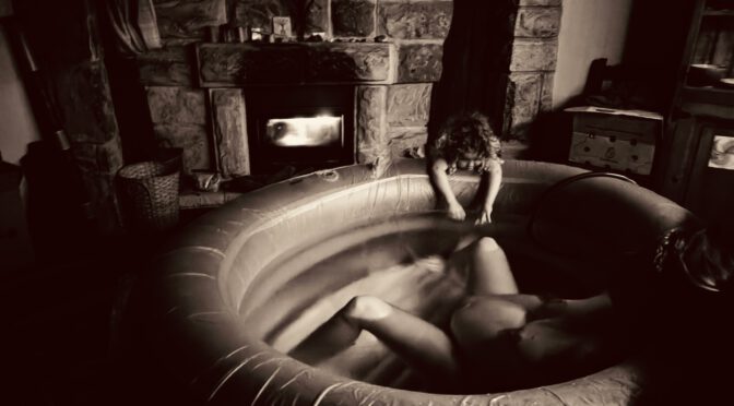 Eine Schwangere liegt im warmen Wasser im Geburtspool, im Hintergrund flackert das Feuer im Kamin und über dem Kaminsims hängen Affirmationskarten zur Motivation: Alleingeburt - ich kann!